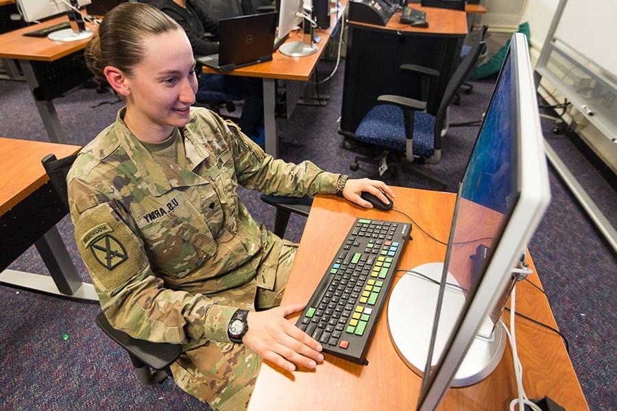 Student in fatigues smiles while working on computer inside.